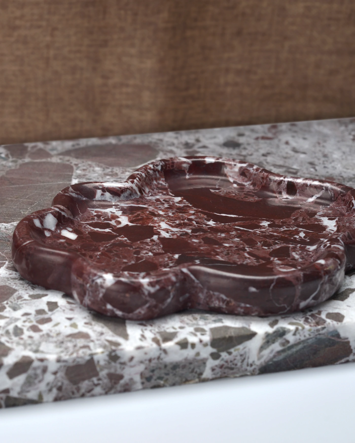 The Cloud Irregular Burgundy Marble Tray with a polished surface, placed on raw marble, featuring red and white veining and scalloped edges