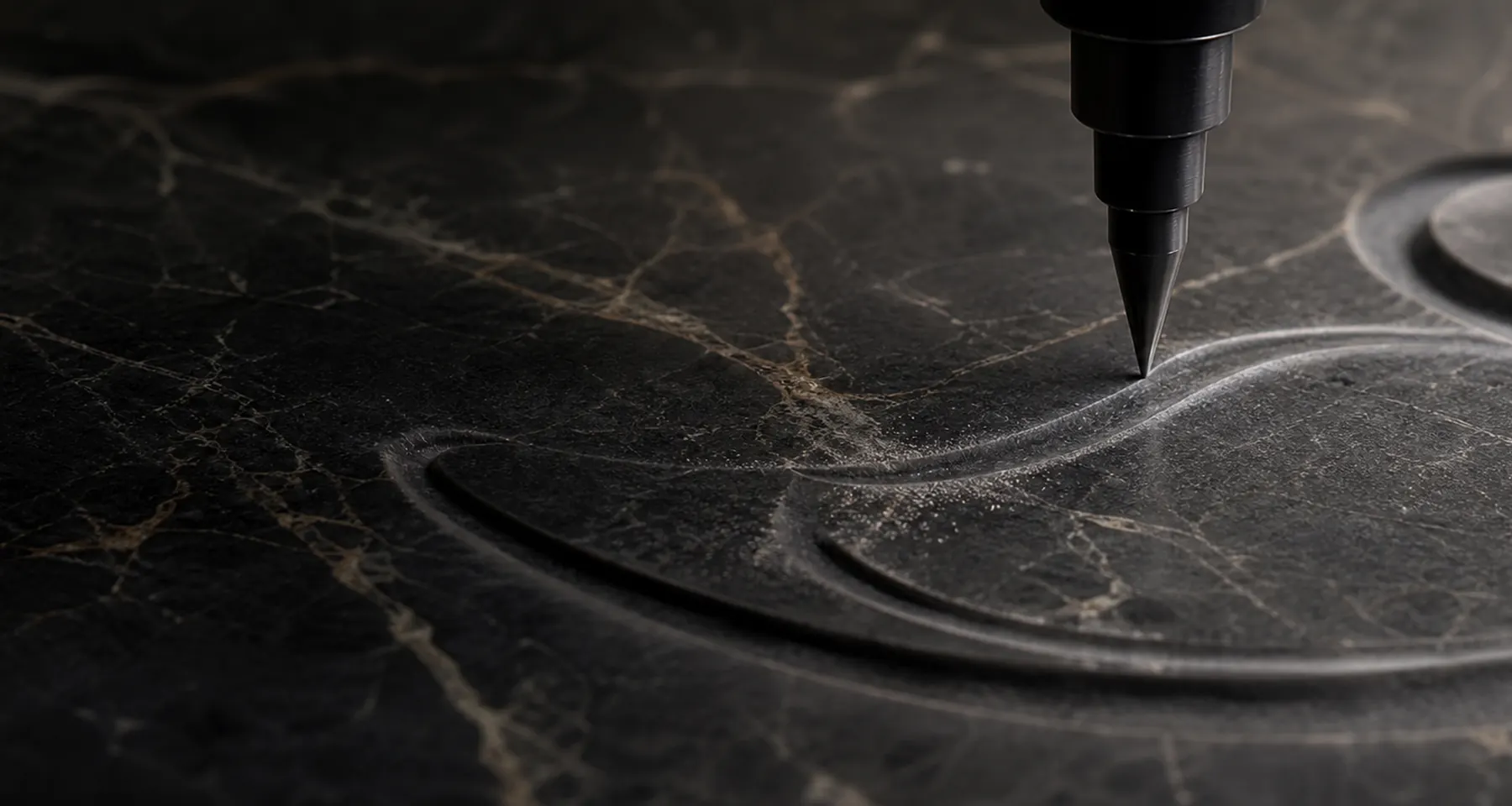 Precision engraving tool carving an abstract groove into dark natural marble.
